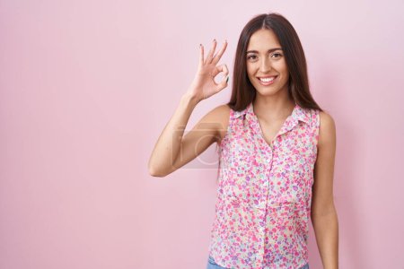 Foto de Mujer hispana joven con el pelo largo de pie sobre fondo rosa sonriendo positiva haciendo signo de ok con la mano y los dedos. expresión exitosa. - Imagen libre de derechos