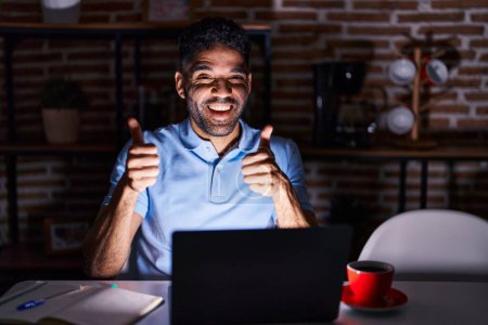 Foto de Hombre hispano con barba usando el ordenador portátil en la noche signo de éxito haciendo gesto positivo con la mano, pulgares hacia arriba sonriendo y feliz. expresión alegre y gesto ganador. - Imagen libre de derechos