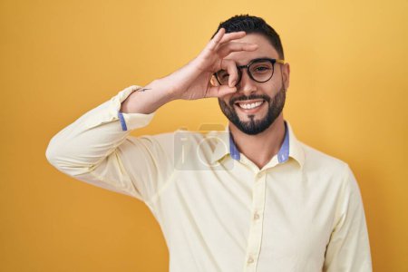 Téléchargez les photos : Jeune homme hispanique portant des vêtements de travail et des lunettes faisant un geste correct avec la main souriante, les yeux regardant à travers les doigts avec un visage heureux. - en image libre de droit