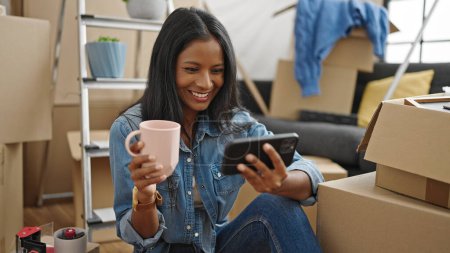 Photo for African american woman watching video on smartphone drinking coffee at new home - Royalty Free Image