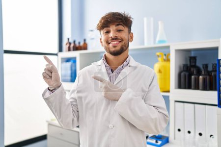 Téléchargez les photos : Homme arabe avec barbe travaillant au laboratoire scientifique souriant et regardant la caméra pointant avec deux mains et doigts sur le côté. - en image libre de droit