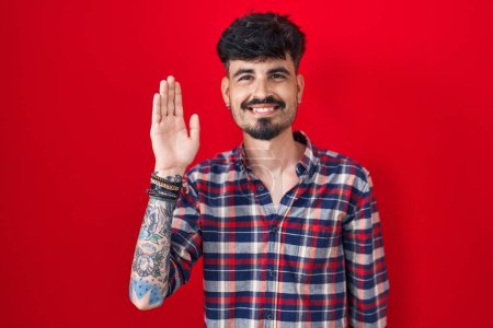 Foto de Joven hombre hispano con barba de pie sobre fondo rojo renunciando a decir hola feliz y sonriente, gesto de bienvenida amistoso - Imagen libre de derechos