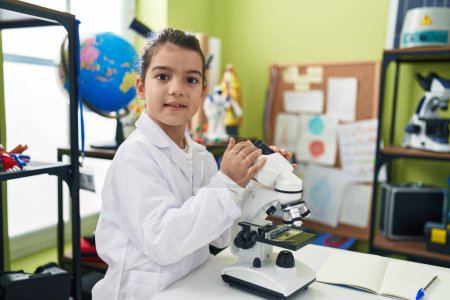 Foto de Adorable estudiante hispana sonriendo confiada usando microscopio en el aula de laboratorio - Imagen libre de derechos