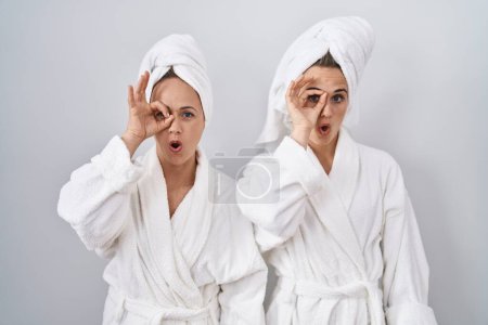 Téléchargez les photos : Femme et fille d'âge moyen portant un peignoir blanc et une serviette faisant un geste correct choqué par le visage surpris, les yeux regardant à travers les doigts. expression incrédule. - en image libre de droit