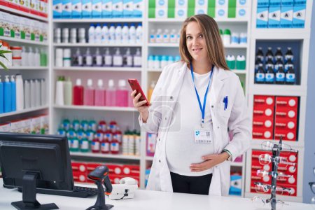 Téléchargez les photos : Jeune pharmacienne enceinte utilisant un smartphone travaillant à la pharmacie - en image libre de droit