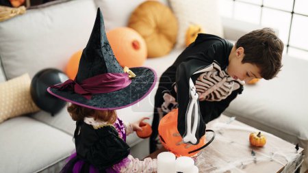Foto de Adorable chico y chica teniendo fiesta de halloween poniendo caramelos en cesta de calabaza en casa - Imagen libre de derechos