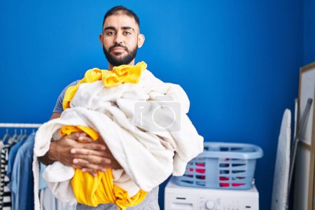 Photo for Middle east man with beard holding pile of laundry relaxed with serious expression on face. simple and natural looking at the camera. - Royalty Free Image