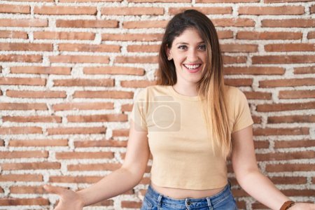Foto de Joven morena de pie sobre la pared de ladrillos sonriente alegre con los brazos abiertos como bienvenida amistosa, saludos positivos y seguros - Imagen libre de derechos