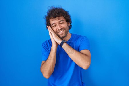 Photo for Hispanic young man standing over blue background sleeping tired dreaming and posing with hands together while smiling with closed eyes. - Royalty Free Image