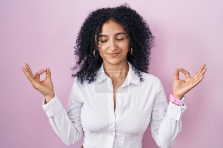 Téléchargez les photos : Femme hispanique avec les cheveux bouclés debout sur fond rose détendue et souriante avec les yeux fermés faisant geste de méditation avec les doigts. concept de yoga. - en image libre de droit
