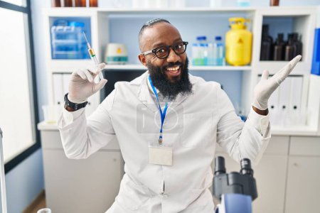 Photo for African american man working at scientist laboratory holding syringe celebrating victory with happy smile and winner expression with raised hands - Royalty Free Image