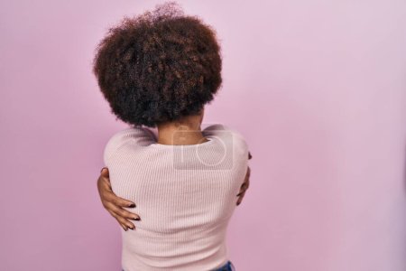 Foto de Joven mujer afroamericana de pie sobre fondo rosa abrazándose feliz y positivo desde atrás. amor propio y cuidado personal - Imagen libre de derechos
