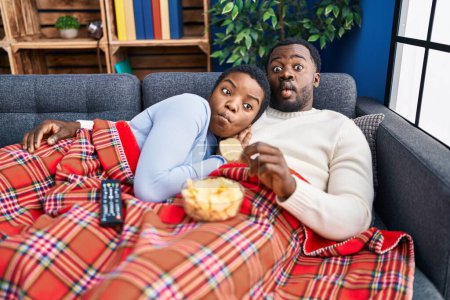 Téléchargez les photos : Jeune couple afro-américain assis sur le canapé regarder la télévision faire visage de poisson avec la bouche et les yeux plissés, fou et comique. - en image libre de droit