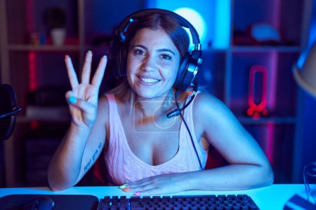 Photo for Young blonde woman playing video games wearing headphones showing and pointing up with fingers number three while smiling confident and happy. - Royalty Free Image