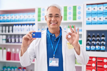 Téléchargez les photos : Homme d'âge moyen avec les cheveux gris travaillant à la pharmacie drugstore tenant carte de crédit faisant signe ok avec les doigts, sourire geste amical excellent symbole - en image libre de droit