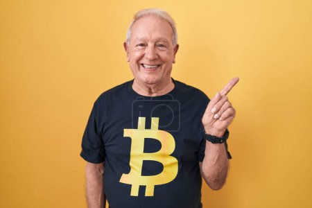 Téléchargez les photos : Homme âgé avec les cheveux gris portant Bitcoin t-shirt avec un grand sourire sur le visage, pointant avec le doigt de la main vers le côté regardant la caméra. - en image libre de droit