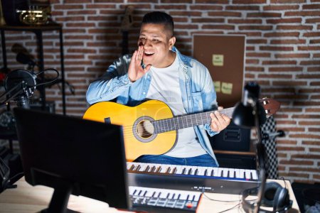 Foto de Joven hispano tocando guitarra clásica en el estudio de música contando rumores secretos, susurrando conversación maliciosa - Imagen libre de derechos
