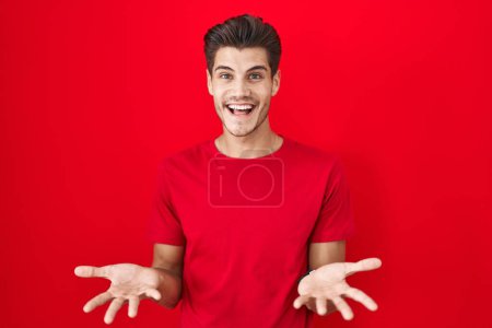 Foto de Joven hombre hispano de pie sobre fondo rojo sonriente alegre con los brazos abiertos como bienvenida amistosa, saludos positivos y confiados - Imagen libre de derechos