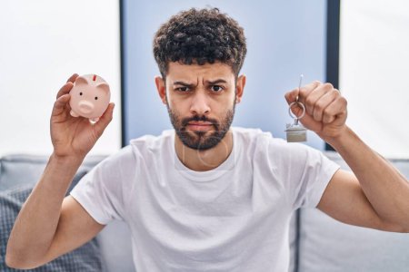 Photo for Arab man holding piggy bank and house keys skeptic and nervous, frowning upset because of problem. negative person. - Royalty Free Image