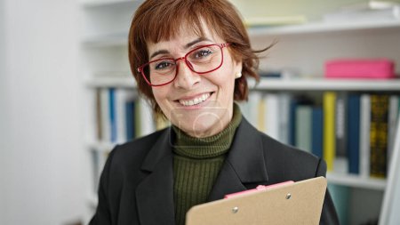 Photo for Mature hispanic woman teacher standing by white board with clipboard at library university - Royalty Free Image