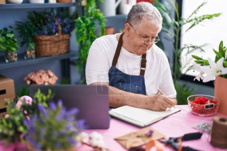 Photo for Middle age grey-haired man florist writing on notebook using laptop at florist - Royalty Free Image