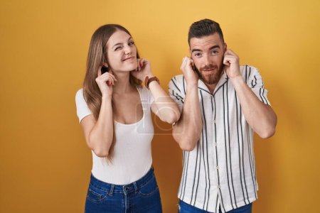 Téléchargez les photos : Jeune couple debout sur fond jaune couvrant les oreilles avec des doigts avec une expression agacée pour le bruit de la musique forte. concept sourd. - en image libre de droit