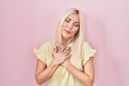 Photo for Caucasian woman standing over pink background smiling with hands on chest with closed eyes and grateful gesture on face. health concept. - Royalty Free Image