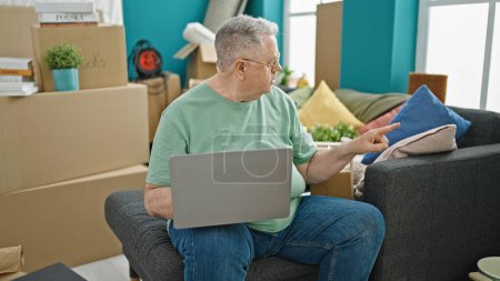 Foto de Hombre de pelo gris de mediana edad usando un portátil sentado en un sofá en un nuevo hogar - Imagen libre de derechos