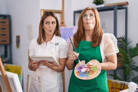 Téléchargez les photos : Peinture hispanique mère et fille au studio de peinture faire visage de poisson avec la bouche et les yeux plissés, fou et comique. - en image libre de droit