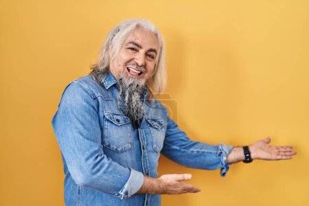 Foto de Hombre de mediana edad con el pelo gris de pie sobre el fondo amarillo invitando a entrar sonriendo natural con la mano abierta - Imagen libre de derechos