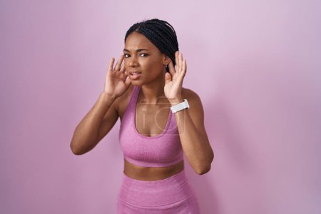 Foto de Mujer afroamericana con trenzas que usan ropa deportiva sobre fondo rosa tratando de escuchar ambos gestos de las manos en la oreja, curioso por chismes. problema auditivo, sordo - Imagen libre de derechos