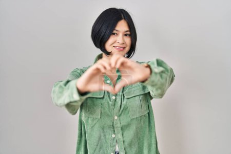 Téléchargez les photos : Jeune femme asiatique debout sur fond blanc souriant dans l'amour faisant forme de symbole de coeur avec les mains. concept romantique. - en image libre de droit