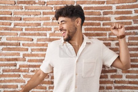 Téléchargez les photos : Homme arabe avec barbe debout sur des briques fond de mur dansant heureux et joyeux, souriant décontracté et confiant écoute de la musique - en image libre de droit