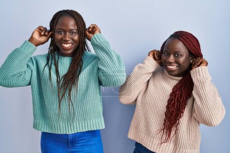 Téléchargez les photos : Deux femmes africaines debout sur fond bleu souriant tirant des oreilles avec les doigts, geste drôle. problème d'audition - en image libre de droit