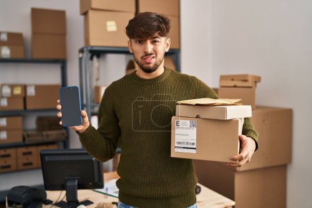 Foto de Hombre árabe con barba trabajando en el comercio electrónico de pequeñas empresas sosteniendo paquetes de entrega sin pistas y expresión confusa. concepto de duda. - Imagen libre de derechos