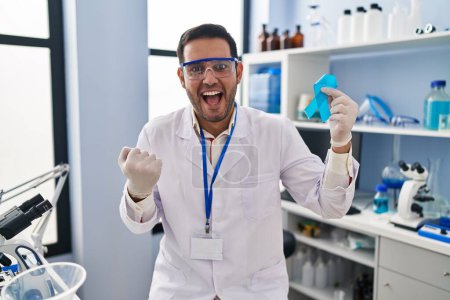 Téléchargez les photos : Jeune homme hispanique avec barbe travaillant au laboratoire scientifique tenant un ruban bleu criant fier, célébrant la victoire et le succès très excité avec les bras levés - en image libre de droit