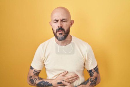 Téléchargez les photos : Homme hispanique avec des tatouages debout sur fond jaune avec la main sur le ventre parce que l'indigestion, la maladie douloureuse se sent mal. concept de douleur. - en image libre de droit