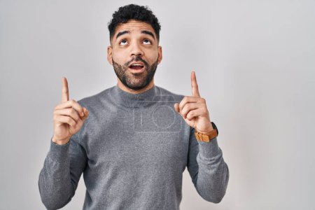 Foto de Hispanic man with beard standing over white background amazed and surprised looking up and pointing with fingers and raised arms. - Imagen libre de derechos