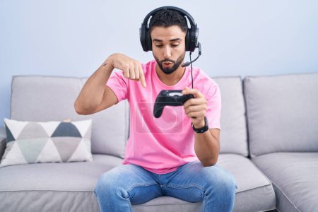 Photo for Hispanic young man playing video game holding controller sitting on the sofa pointing down with fingers showing advertisement, surprised face and open mouth - Royalty Free Image