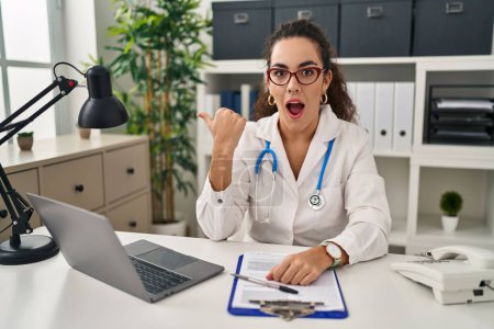 Photo for Young hispanic woman wearing doctor uniform and stethoscope surprised pointing with hand finger to the side, open mouth amazed expression. - Royalty Free Image