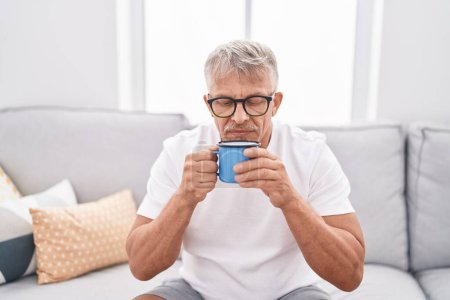Foto de Hombre de pelo gris de mediana edad bebiendo café sentado en el sofá en casa - Imagen libre de derechos