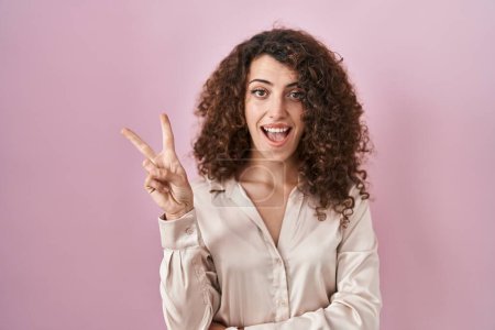 Photo for Hispanic woman with curly hair standing over pink background smiling with happy face winking at the camera doing victory sign with fingers. number two. - Royalty Free Image
