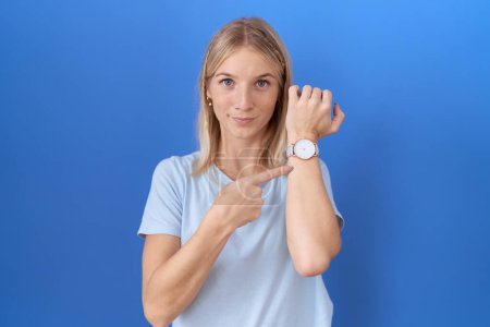 Téléchargez les photos : Jeune femme caucasienne vêtue de t-shirt bleu décontracté à la hâte pointant vers le temps de montre, impatience, regardant la caméra avec une expression détendue - en image libre de droit