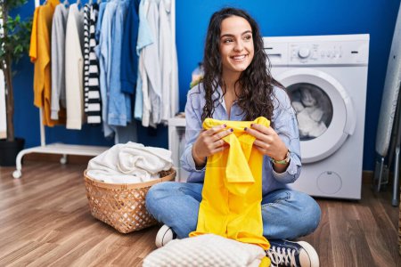 Téléchargez les photos : Jeune femme hispanique pliant vêtements en attente de machine à laver à la buanderie - en image libre de droit