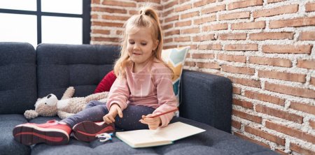 Foto de Adorable chica rubia estudiante de preescolar sentado en el sofá dibujo en el cuaderno en casa - Imagen libre de derechos