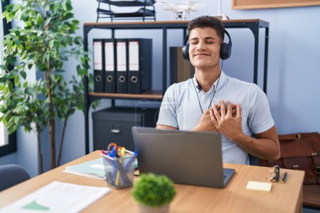 Téléchargez les photos : Jeune homme hispanique travaillant au bureau portant des écouteurs souriant avec les mains sur la poitrine avec les yeux fermés et geste reconnaissant sur le visage. concept de santé. - en image libre de droit