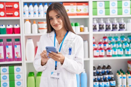 Photo for Young beautiful hispanic woman pharmacist using smartphone working at pharmacy - Royalty Free Image