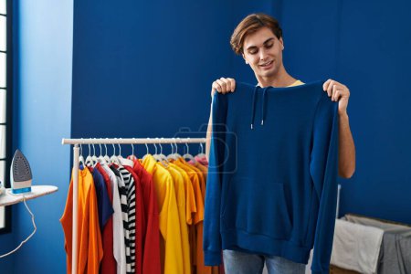 Téléchargez les photos : Young caucasian man smiling confident holding sweatshirt at laundry room - en image libre de droit
