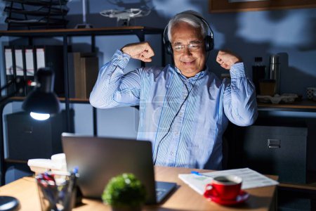 Photo for Hispanic senior man wearing call center agent headset at night showing arms muscles smiling proud. fitness concept. - Royalty Free Image