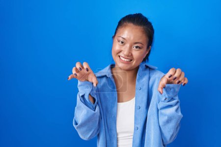 Téléchargez les photos : Asiatique jeune femme debout sur fond bleu souriant drôle faisant griffe geste comme chat, expression agressive et sexy - en image libre de droit